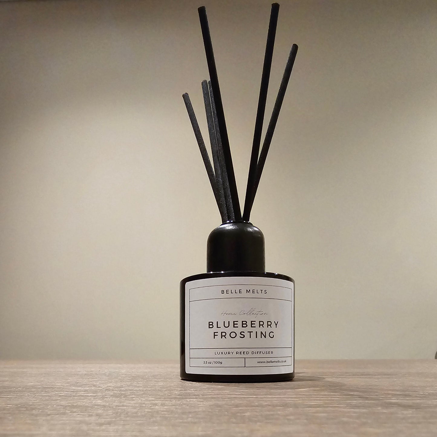 Bottle of 'Blueberry Frosting' diffuser with black sticks on a wooden surface and beige background