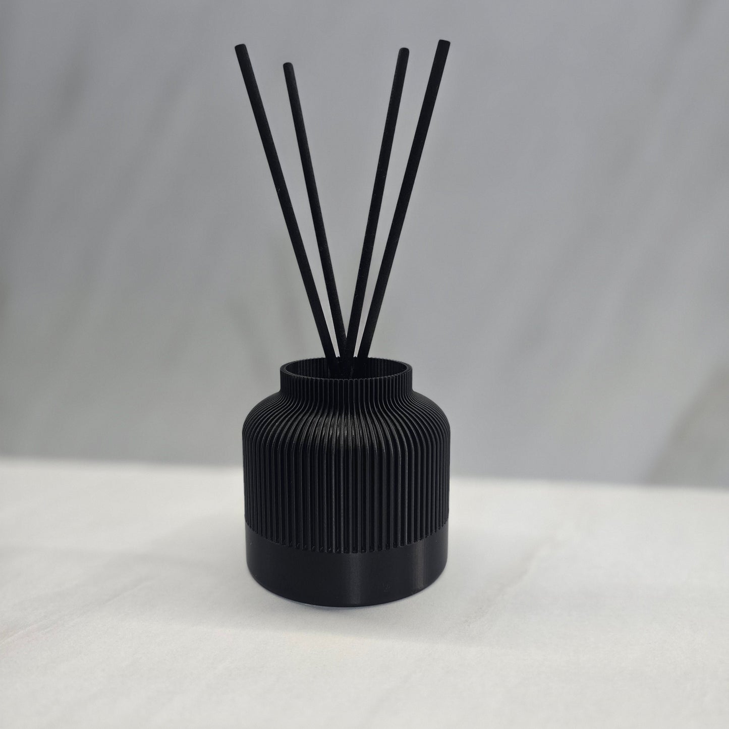 A black reed diffuser with several black reeds inserted into a textured, 3D printed black vessel on a light background.