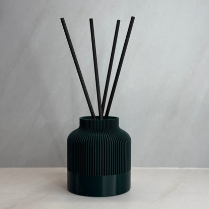 A black reed diffuser with several black reeds inserted into a textured, 3D printed black vessel on a light background.