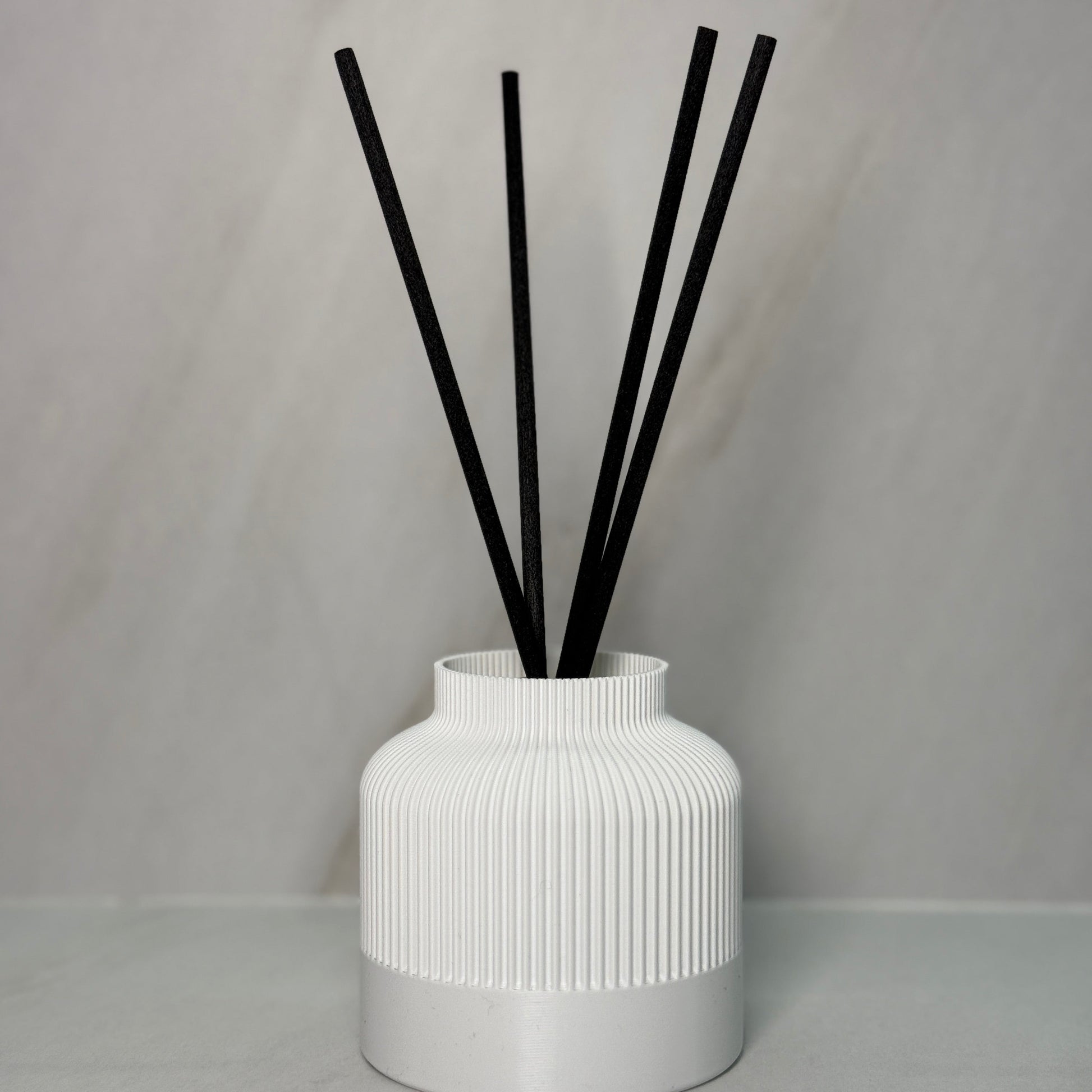 A black reed diffuser with several black reeds inserted into a textured, 3D printed black vessel on a light background.