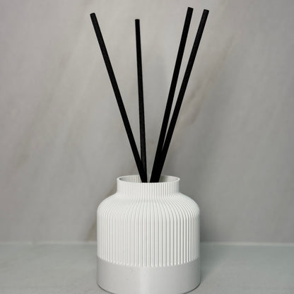A black reed diffuser with several black reeds inserted into a textured, 3D printed black vessel on a light background.