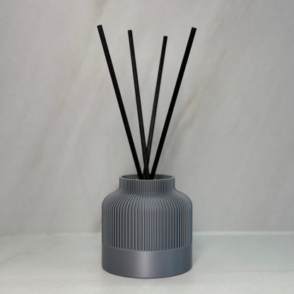 A black reed diffuser with several black reeds inserted into a textured, 3D printed black vessel on a light background.