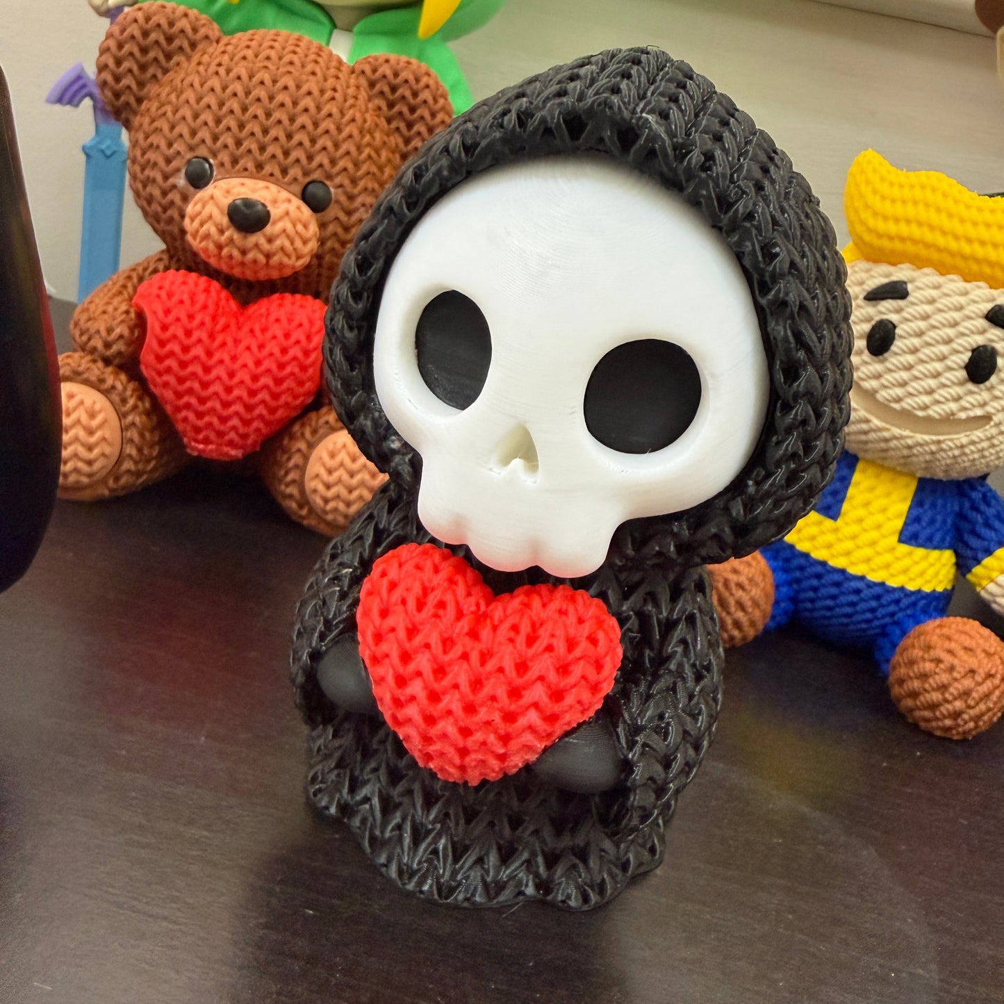 Crocheted skull with red heart surrounded by small figurines on a reflective surface