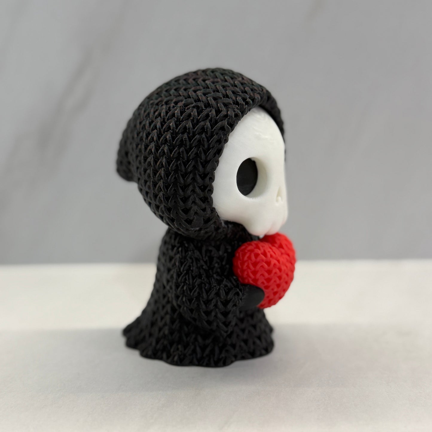 Knitted toy resembling a character with a black hood and red heart on a white background